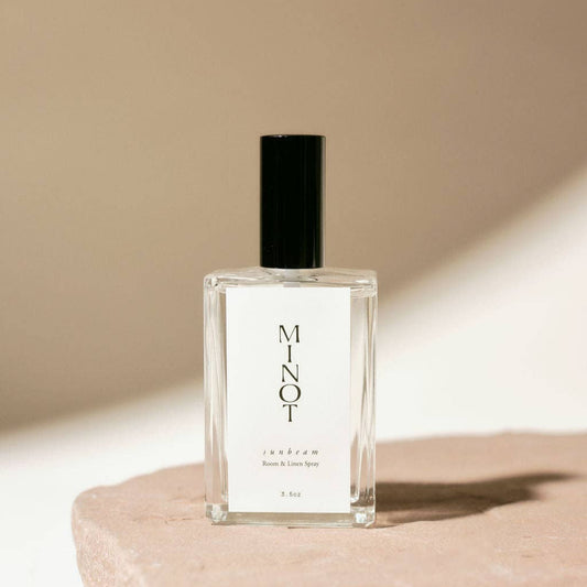 A clear glass bottle with a black spray nozzle, labeled 'MINOT', containing a room & linen spray with a quantity of over 1,000 sprays, displayed on a beige surface.