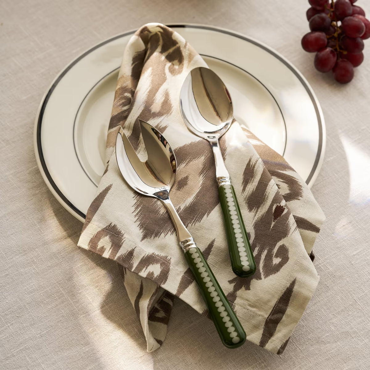 Two silver spoons with dark green handles featuring white dotted accents, arranged on a beige and brown patterned cloth napkin over a white plate with a black rim, on a beige tablecloth with a bunch of red grapes in the corner.