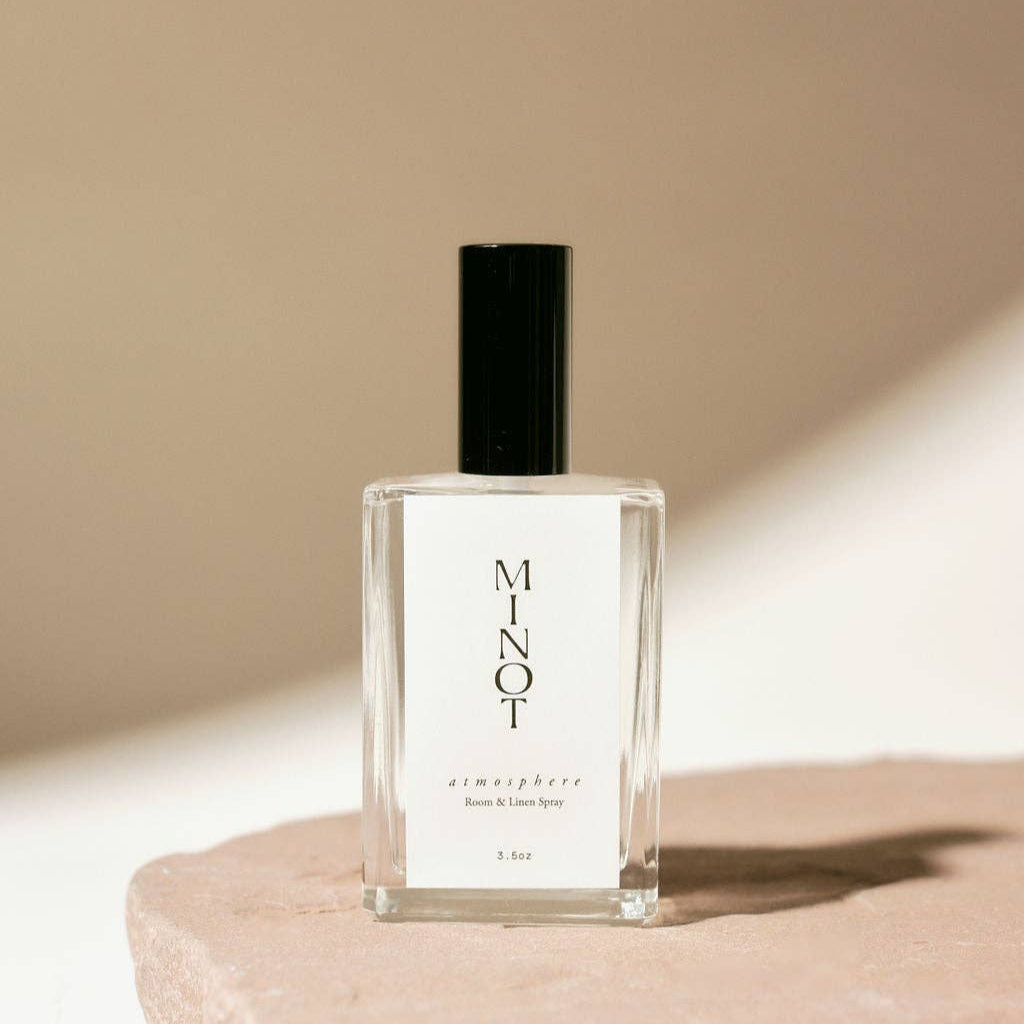 A clear glass bottle of Atmosphere Room & Linen Spray with a black cap, labeled with the brand name and scent information, placed on a beige surface.